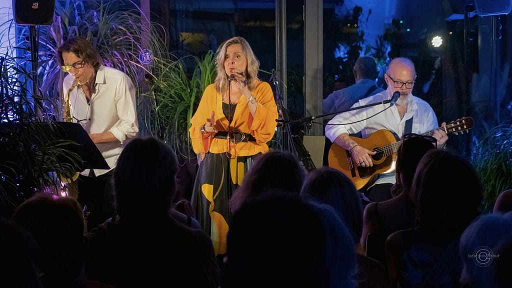 Musiciens sur scène interprétant un concert acoustique brésilien avec chant, guitare et saxophone.